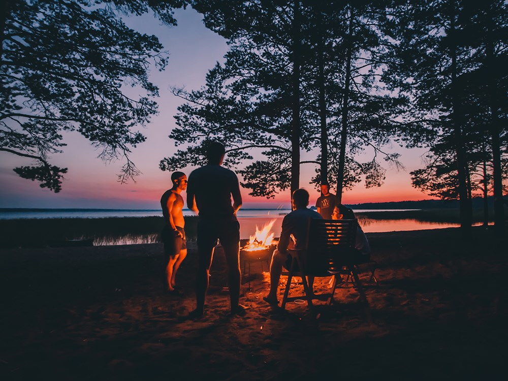 Group of young people near camp fire with campfire song and campfire meals playing campfire games and eating camp fire grill, telling campfire stories near the fire with wood, flames in the nature at night near lake.