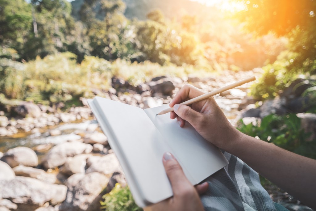 Woman hand writing on notebook with pencil at river background.